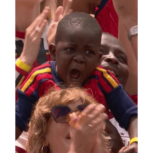 Niño pequeño con la boca abierta y ojos muy abiertos, vestido con una camiseta de colores rojo, amarillo y azul. Una mujer con peluca rubia y gafas de sol lo so